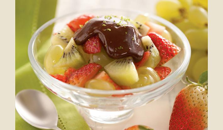 fotografia em tons de verde, contém uma taça transparente com pedaços de morangos, kiwis e ganache por cima