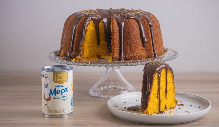 Foto da receita de Bolo de Cenoura com Cobertura de Brigadeiro, servido em um prato alto de vidro, com um pedaço cortado ao lado e, do lado esquerdo, uma lata de Leite Moça