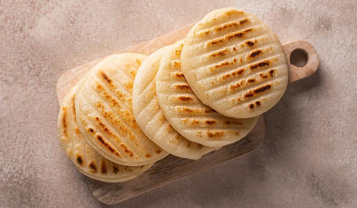 Fotografia vista de cima em tons claros de cinco arepas médias sobrepostas apoiadas em uma mini tábua retangular de madeira, sobre uma mesa clara.
