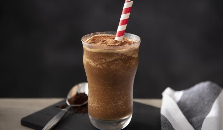 Fotografia em tons de marrom em uma bancada de madeira de cor cinza. Ao centro, uma tábua de madeira preta contendo um copo em cima com o shake. Ao lado, há uma colher colher com nescafé e um pano listrado cinza e branco.