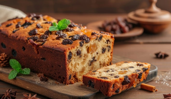 Fotografia de um bolo de frutas recém-assado, com crosta dourada e rica em passas e frutas secas. A fatia cortada revela a textura úmida, em um ambiente rústico com elementos decorativos como anis estrelado e canela.