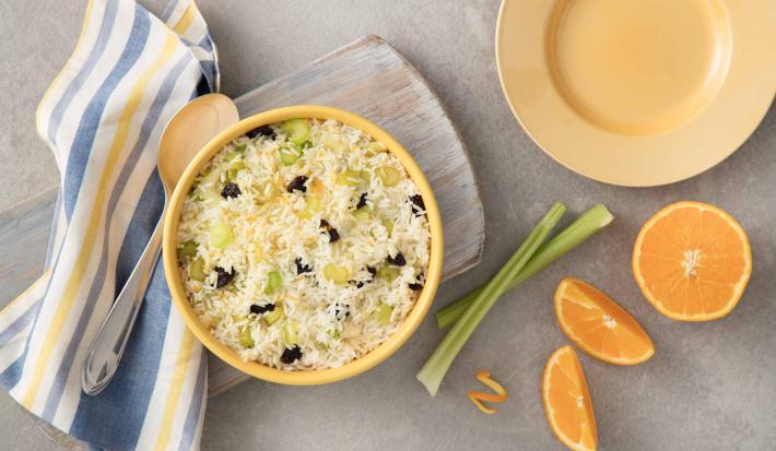 Fotografia em tons de azul e laranja em uma bancada de madeira cinza, um pano listrado amarelo e azul, uma tábua de madeira mesclada com um pote redondo grande amarelo com o arroz com laranja dentro dele. Ao lado, alho-poró e gomos de laranja.