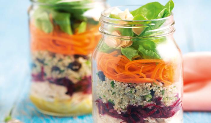 Fotografia em tons de azul em uma bancada de madeira azul, dois potinhos de vidro com a salada de quinoa, beterraba, cenoura e agrião dentro deles. Ao lado, um paninho rosa e dois garfos.