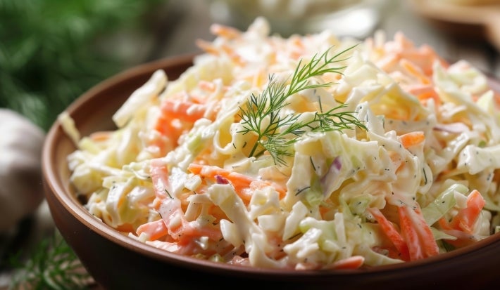 Em cima de uma mesa,  tigela com salada de maionese cremosa e colorida com cenouras raladas e repolho ralado, decoradas com folha verde.