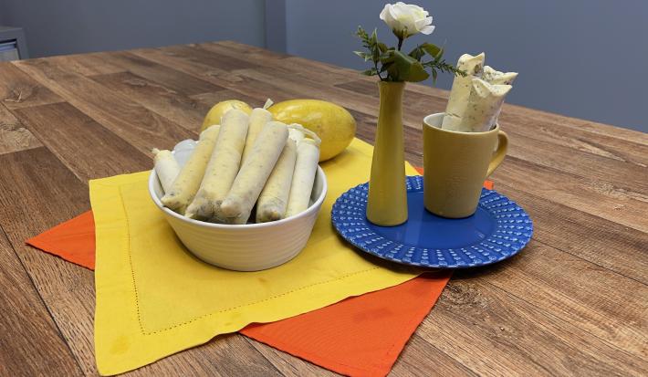 Foto da receita de Sacolé de Maracujá, servido em saquinhos, em tom amarelo, em uma travessa branca, sobre uma bancada de madeira decora com um tecido amarelo e outro laranja, ao lado há um prato azul redondo com um vaso e uma caneca amarelos