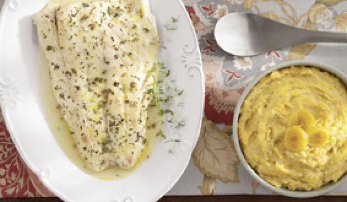 Fotografia em tons de amarelo em uma bancada de madeira com uma toalha florida, um prato oval branco com o peixe temperado com tomilho. Ao lado, um potinho branco com o purê de banana.