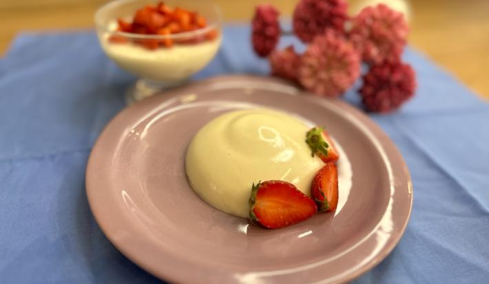 Fotografia em tons de rosa e azul com um prato rosa ao centro. Em cima do prato existe uma receita de Panna Cotta com morangos fatiados por cima