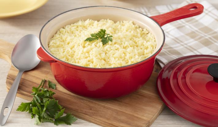 Fotografia em tons de vermelho de uma bancada branca, ao centro uma panela vermelha por fora e banca por dentro com arroz levemente amarelado. No topo, uma folha de salsinha decorando.