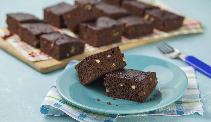 fotografia em tons de azul e marrom de uma bancada vista de frente, contém um pano em tons de branco e azul, um prato azul e redondo contém 2 Brownies e ao fundo um tábua de madeira com diversos Brownies