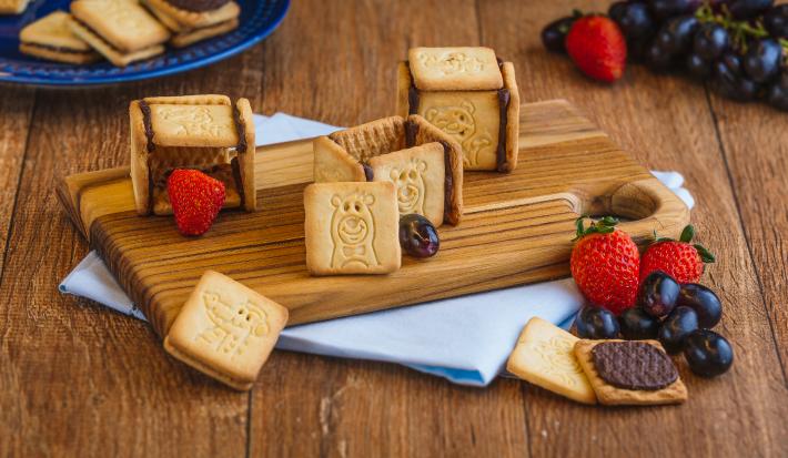 Foto de um balcão de madeira com uma pequena tábua com biscoitos em cima, alguns abertos, alguns morangos e uvas e um tecido em tom azul claro.