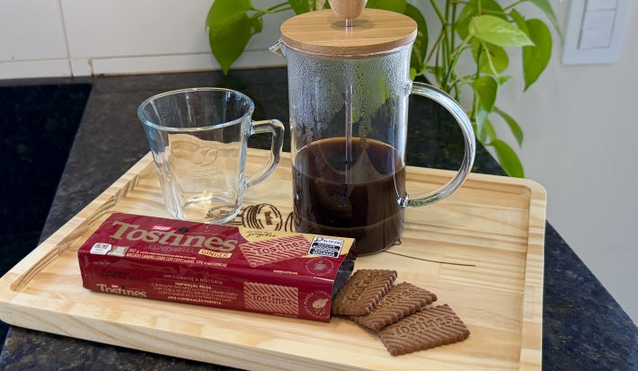Foto da receita de Nescafé Gold com Especiarias, servido em uma prensa francesa, sobre uma bandeja de madeira e com um pacote de Biscoito Tostines Ginger na frente