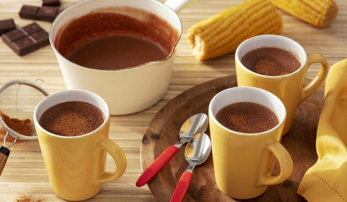 Fotografia em tons de amarelo em uma bancada de madeira de cor marrom. Ao centro, uma bandeja de madeira contendo 2 xícaras amarelas com 2 colheres ao lado. Ao fundo, há 2 espigas de milho, uma panela branca e uma xícara com chocolate quente.