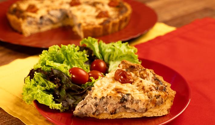 Fotografia mostra um pedaço de quiche cortada sobre um prato vermelho, ao lado folhas verdes de alface e tomate cereja.