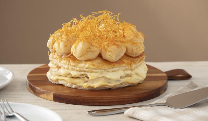 Foto da Receita de Gateau Saint Honoré. Observa-se a torta folhada sobre uma tábua redonda de madeira, num fundo e sobre uma superfície cor nude. Na ambientação, tem-se uma espátula de prata e um guardanapo quadriculado.