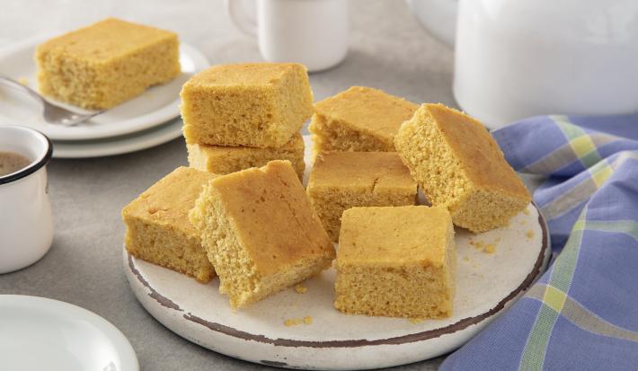 Foto da receita de Bolo de fubá sem glúten. Observa-se um prato de cerâmica com fatias quadradas do bolo e, ao lado esquerdo, uma xícara branca de café.