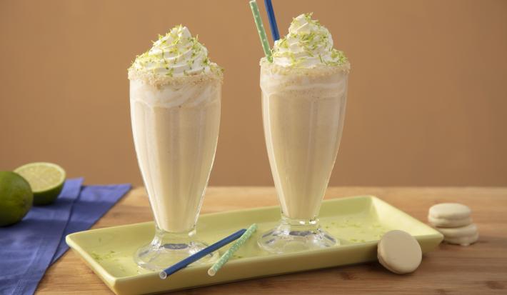 Foto da receita de Milkshake de Bono Torta de Limão Coberto, em dois copos decorados com chantilly, sobre um prato verde, em uma bancada de madeira decora com fatias de limão, biscoitos brancos e um tecido em tom azul
