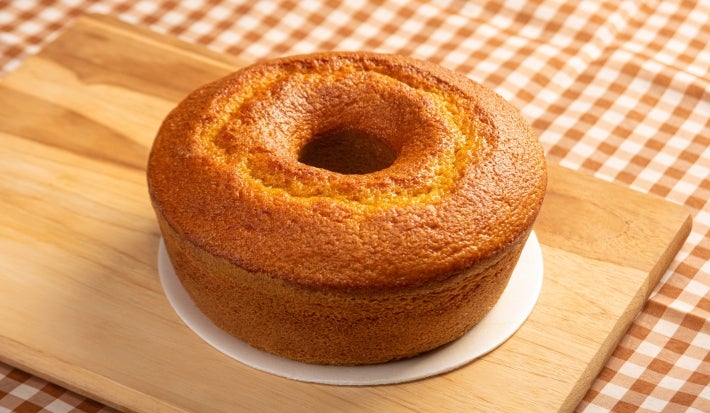 O bolo de cenoura simples com furo no meio e aparência bem fofinha. Ele está em uma base branca sobre uma tábua de madeira, com uma toalha de mesa xadrez ao fundo, criando um clima caseiro e acolhedor.