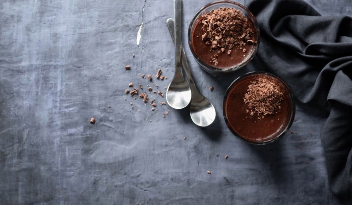 Fotografia vista de cima em tons escuros de dois copos de vidro com a sobremesa, finalizada com a ganache e raspas de chocolate. Ao redor tem duas colheres de sobremesa e um pano dobrado preto, sobre uma mesa de tom de cimento queimado escuro.