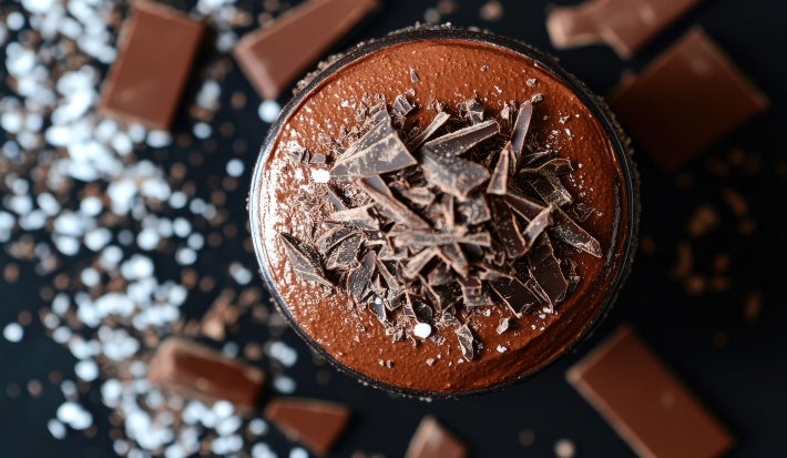 Fotografia em tons mais escuros, vista de cima, de um pote de vidro com a sobremesa, finalizada com ganache e raspas de chocolate. O pote está sobre uma mesa preta com pedaços de chocolate.