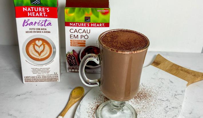 Foto da receita de chocolate quente cremoso servida em uma xícara alta e larga de vidro sobre uma tábua de mármore branca. Ao fundo há duas caixas de natures heart, uma de bebida vegetal barista e outra de cacau em pó