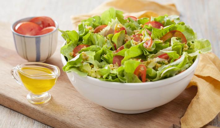 Fotografia em tons de  marrom e branco de uma bancada de madeira com uma tábua de madeira, sobre ela um recipiente branco redondo com salada. Ao lado um paninho amarelo, um recipiente com azeite e um recipiente com tomate.