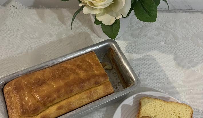 Fotografia vista de cima de um pão dentro de uma forma pequena retangular de alumínio ao lado de um prato pequeno raso com uma fatia do pão.