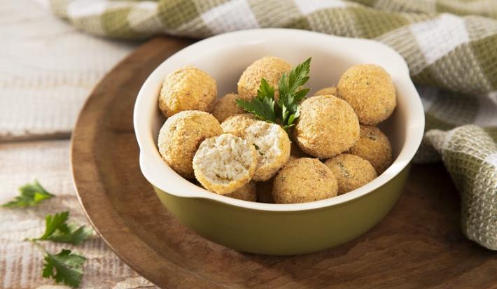 Foto em tons de bege da receita de bolinho proteico de frango servida em diversas porções dentro de uma panelinha de porcelana verde sobre uma tábua circular de madeira com um pano quadriculada verde musgo