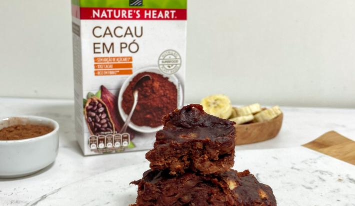Foto em tons de marrom da receita de brownie de 3 ingredientes servida em diversas porções sobrepostas sobre uma de mármore branca. Ao fundo há um bowl com rodelas de banana e um com cacau em pó