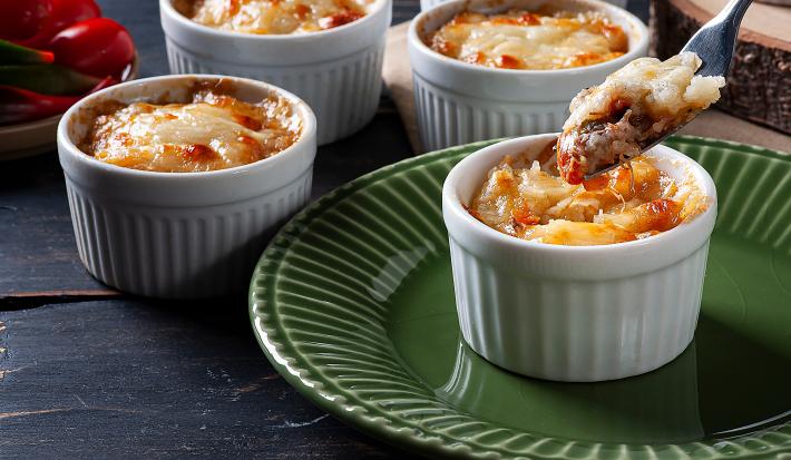 Fotografia em tons de marrom com vários ramekins ao centro. Dentro de cada recipiente existe uma receita de escondidinho de carne seca sem lactose. Um prato de mandiocas gratinadas com carne seca.