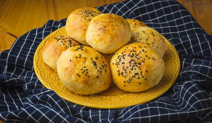 Foto aproximada de um prato amarelo com alguns pães de batata grande, decorados com gergelim. O prato está sobre um tecido azul quadriculado e numa bancada de madeira.