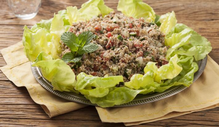 Em uma mesa de madeira contém panos em tons pardo abaixo de um recipiente que comporta a salada para quibe.