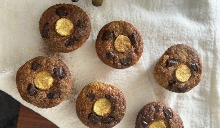 Fotografia em tons de branco com 6 muffins de banana ao centro. Os Muffins são finalizados com canela e uma fatia de banana por cima.