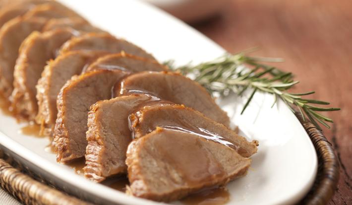 foto em tons marrom e branco. Uma mesa de madeira contém um cesto com um prato que comporta pedaços de carne fatiados.