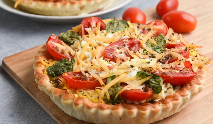 Imagem de uma quiche sobre uma tábua de madeira. A massa da quiche é dourada e crocante e ela está recheada com tomates-cereja, folhas de manjericão e lascas de queijo parmesão.