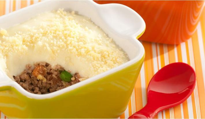 fotografia em tons de amarelo, branco e laranja tirada de uma bancada nas cores branco e laranja. Contém um recipiente redondo amarelo com alças com um purê recheado com carne moída dentro.