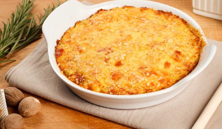 Fotografia de um recipiente refratário de vidro branco sobre um pano cinza sobre uma mesa de madeira clara. Dentro do recipiente tem uma receita de bolo de batata com queijo gratinado por cima.
