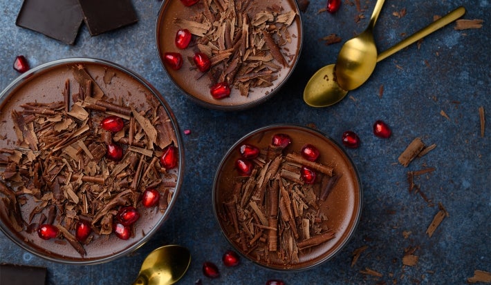 Fotografia em tons escuros, vista de cima, de três recipientes de vidro, grandes, sobre uma mesa de tom escuro, com algumas colheres douradas, raspas e pedaços de chocolate. Dentro dos recipientes tem a sobremesa, finalizada com ganache e raspas.