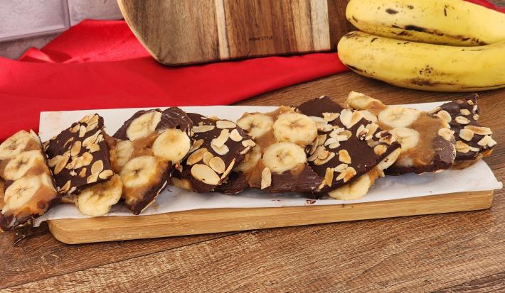 Fotografia em tons de vermelho em uma bancada de madeira com uma tábua e em cima barras de banana com chocolate e amêndoas . Ao fundo, um cacho de banana e um pano vermelho.