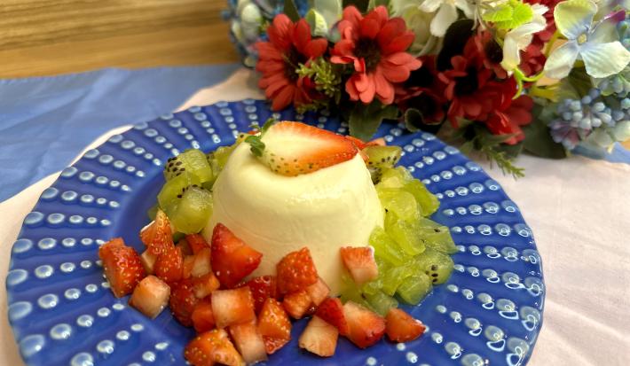 Fotografia em tons de azul com um prato azul ao centro. Em cima do prato existe uma receita de pudim feito com ASCENDA acompanhado de fatias de kiwi e morango