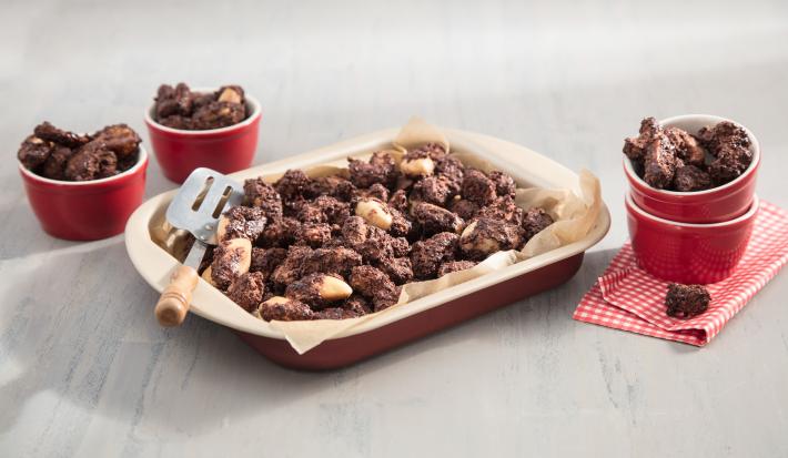 Fotografia em tons de vermelho em uma bancada de madeira clara com um refratário vermelho ao centro com castanhas crocantes com chocolate. Ao lado, potinhos vermelhos com pedaços de castanhas envoltas de chocolate.