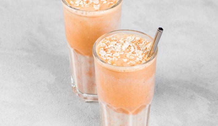 Fotografia de dois copos de vidro com um suco de pitanga com melaço de cana. Dentro dos copos com o suco, um canudo de metal, e embaixo dos copos, uma mesa cinza em tom claro.