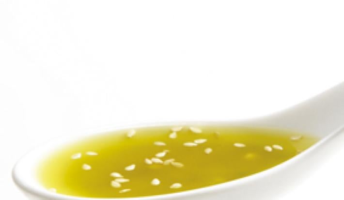 Fotografia em tons de amarelo em um fundo e mesa brancos com uma colher com o molho de mostarda com gergelim.