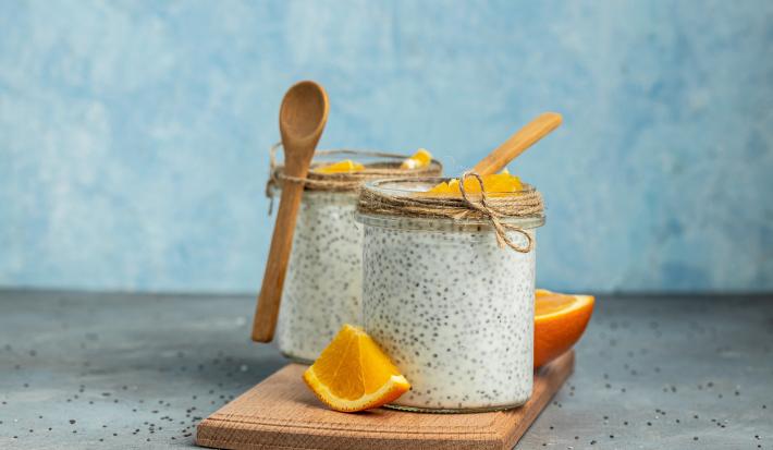 Fotografia em tons de azul com dois copos de vidro ao centro. Dentro de cada pote existe uma receita de Overnight Chia feita com laranja, chia e coco. Ao fundo existe uma laranja cortada ao meio