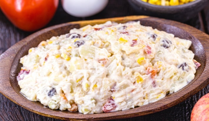 Fotografia de uma salada cremosa em uma tigela de madeira, com batatas, cenouras, ervilhas, uva-passa e milho, destacando a textura rica e ingredientes frescos, como um tomate e um ovo ao fundo, criando um ambiente rústico e acolhedor.
