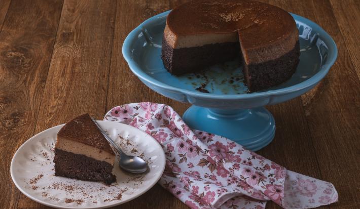 Foto da receita de bolo chocopudim da maria carpio, servido sobre um prato grande azul, com uma fatia cortada sobre outro prato branco, tudo em uma bancada de madeira
