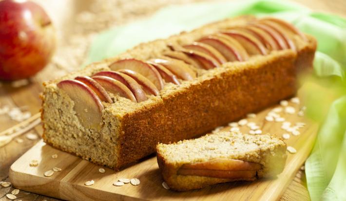 Fotografia em tons de marrom de uma bancada de madeira, sobre ela uma tábua de madeira com o bolo de maça. Ao fundo uma maçã e um paninho verde.