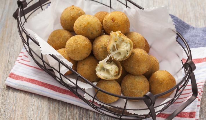 Fotografia em tons cinza, branco e dourado, mostrando bancada em tons de cinza com cestinha de arame contendo bolinhas de queijo fritas sobre papel toalha e guardanapo branco com listras vermelhas.
