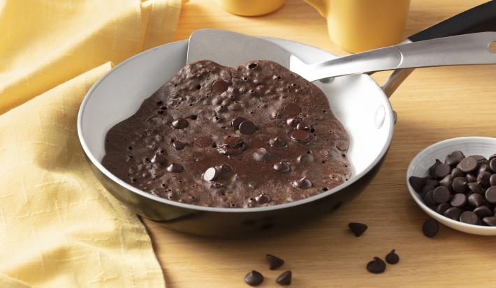 Fotografia em tons de amarelo em uma bancada amarela e um pano amarelo na lateral. Uma frigideira ao centro com o brownie de chocolate dentro dela. Ao lado, um potinho com gotas de chocolate.
