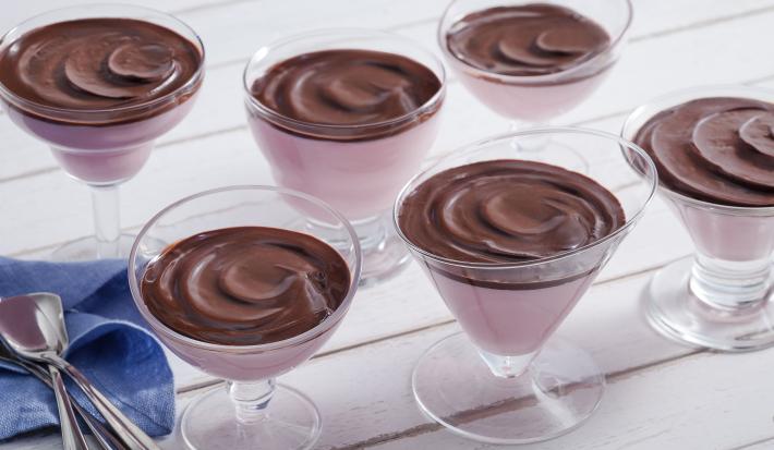 Fotografia em tons de chocolate e rosa, com seis taças contendo creme rosa e creme de chocolate, ao lado guardanapo azul e várias colheres, tudo sobre bancada em tom de rosa.