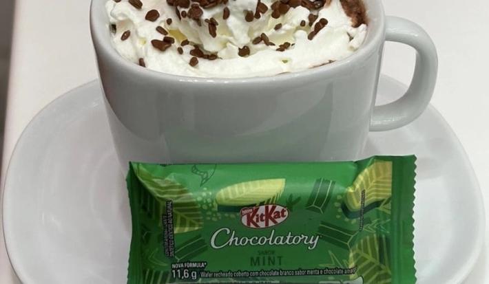 Fotografia de uma xícara branca com um hot chocolate com chantilly e granulado por cima. Na frente da xícara tem um kitkat de menta, e está sobre um prato pequeno de vidro raso branco.